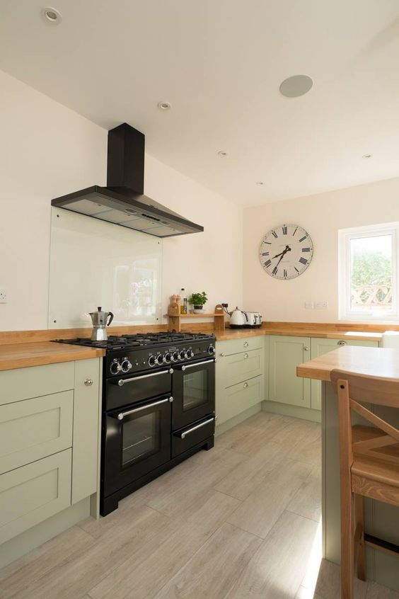 a beautiful sage green kitchen with butcherblock countertops, a black cooker and a hood, a glass backsplash is chic