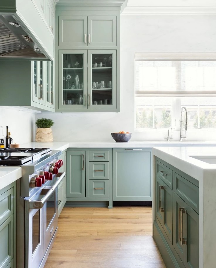 a beautiful sage green kitchen with shaker and glass front cabinets, white stone countertops and a backsplash feels airy