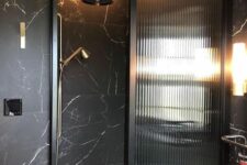 a black bathroom clad with marble, with a fluted glass space divider, brass touches for a more elegant and chic look
