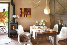 a bright dining space with a stained table, white curved chairs, a pendant lamp, a hearth, a bold artwork and a terracotta tile floor