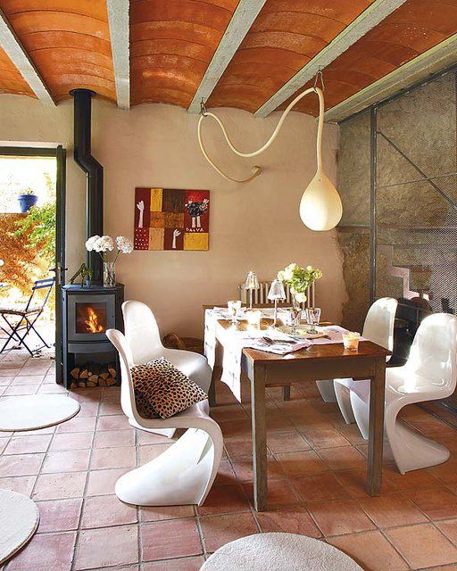 a bright dining space with a stained table, white curved chairs, a pendant lamp, a hearth, a bold artwork and a terracotta tile floor