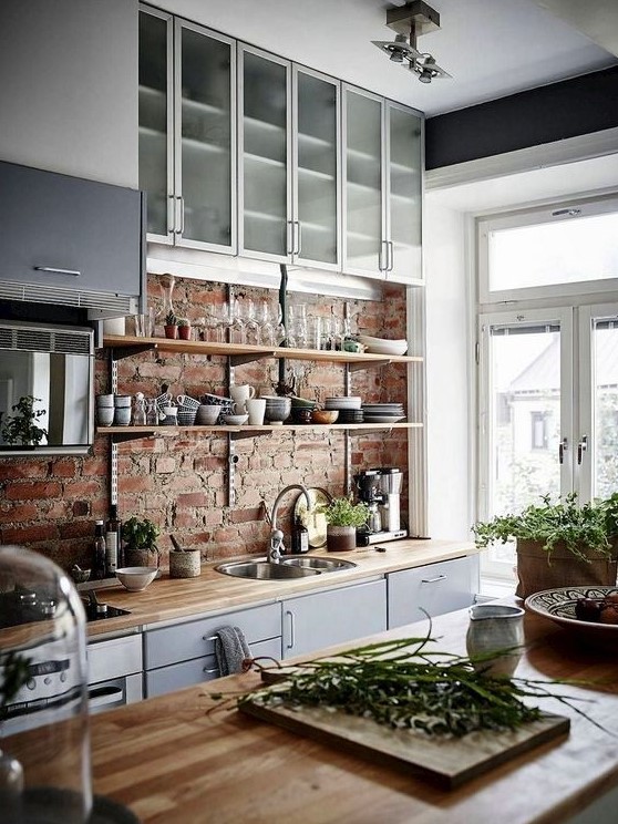 a chic Scandinavian kitchen with a brick wall, grey and frosted glass cabinets, butcherblock countertops and much light