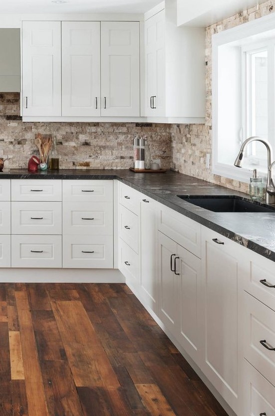 a chic white kitchen with a tan stone tile backsplash and black granite countertops plus black handles is a stylish space