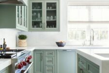 a classic sage green shaker style kitchen with glass front cabinets, white stone countertops and backsplash and brass handles