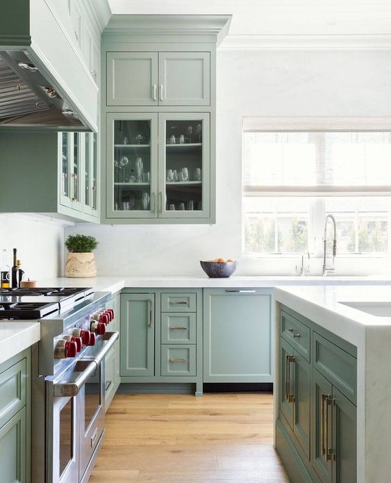 a classic sage green shaker style kitchen with glass front cabinets, white stone countertops and backsplash and brass handles