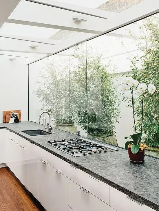 a contemporary white kitchen with a dark countertop, a glazed wall with a view of the private garden and greenery