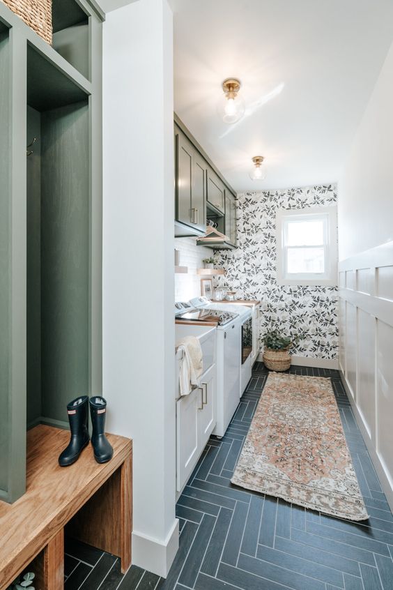 a cool and sleek mudroom entry with a wallpaper accent wall, green and grey cabinets, a herringbone tile floor and a potted plant