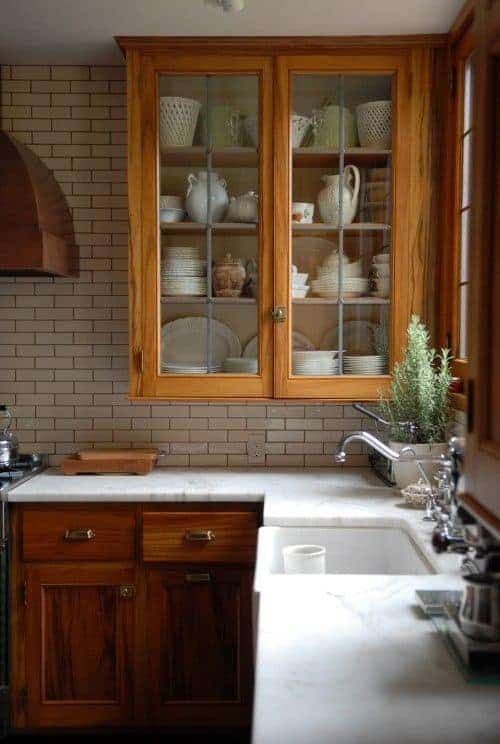 a cozy stained kitchen with shaker and glass front cabinets, a neutral tile backsplash, white quartz countertops and potted plants