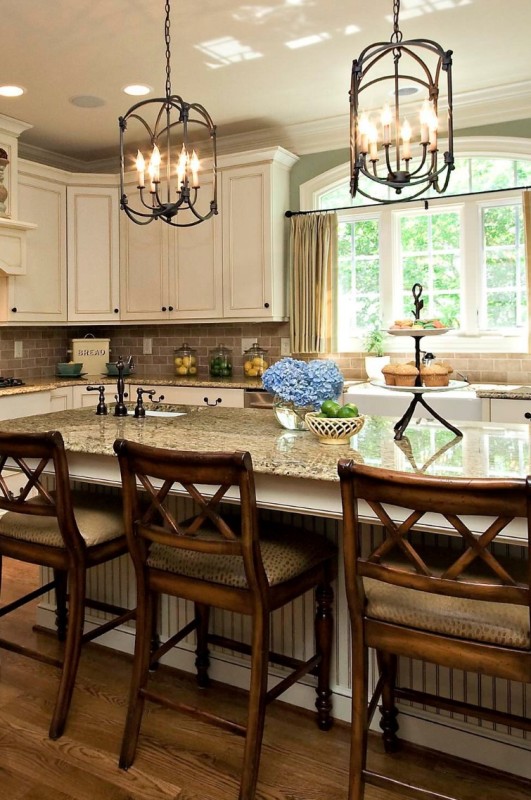 a creamy kitchen with shaker cabinetry, a taupe tile backsplash and beige granite countertops, vintage chandeliers and stained stools