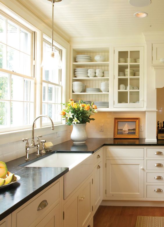 a creamy vintage kitchen with shaker and glass cabinets, black quartz countertops, a window as a backsplash and knobs and handles