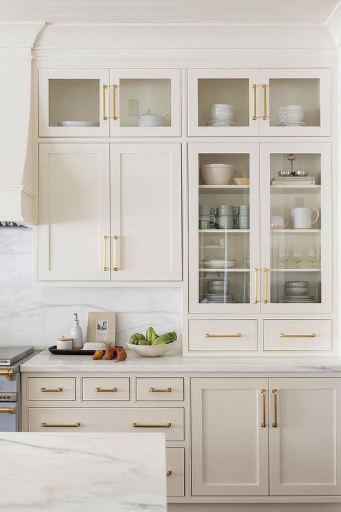 a delicate tan kitchen with shaker and glass front cabinets, gold handles and a white marble backsplash and countertops