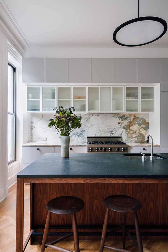 a dove grey flat panel kitchen with glass front cabinets, a white marble backsplash, a stained kitchen island with a black soapstone countertop