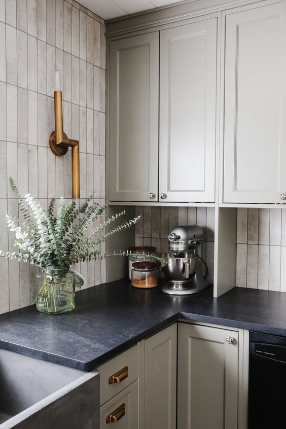 a dove grey shaker style kitchen with a skinny tile backsplash, black soapstone countertops and brass fixtures