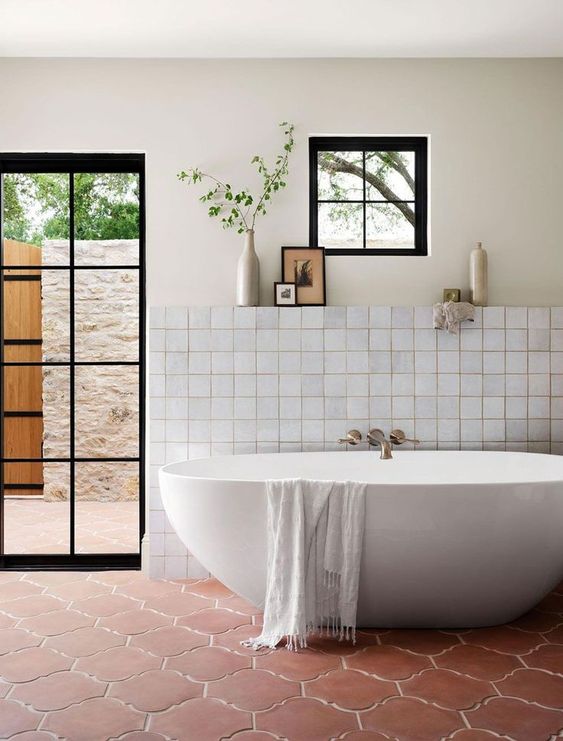 a farmhouse bathroom with neutral tiles, a terracotta tile floor, an oval tub, a glass door to the garden and neutral textiles