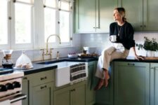 a farmhouse kitchen in sage green, with shaker cabinets, black countertops, a white subway tile backsplash and brass handles