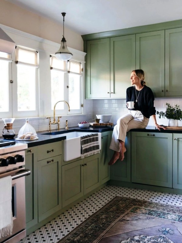 a farmhouse kitchen in sage green, with shaker cabinets, black countertops, a white subway tile backsplash and brass handles