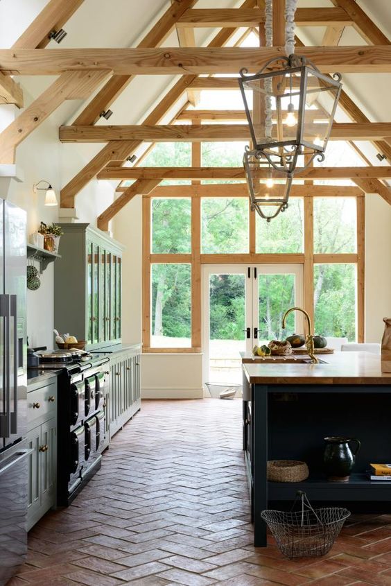 a farmhouse kitchen with a terracotta tile floor, blue cabinets, a navy kitchen island, a black cooker, wooden beams and pendant lamps