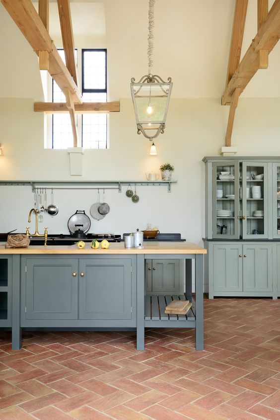 a farmhouse kitchen with a terracotta tile floor, grey cabinetry, a graphite grey kitchen island, wooden beams and pendant lamps