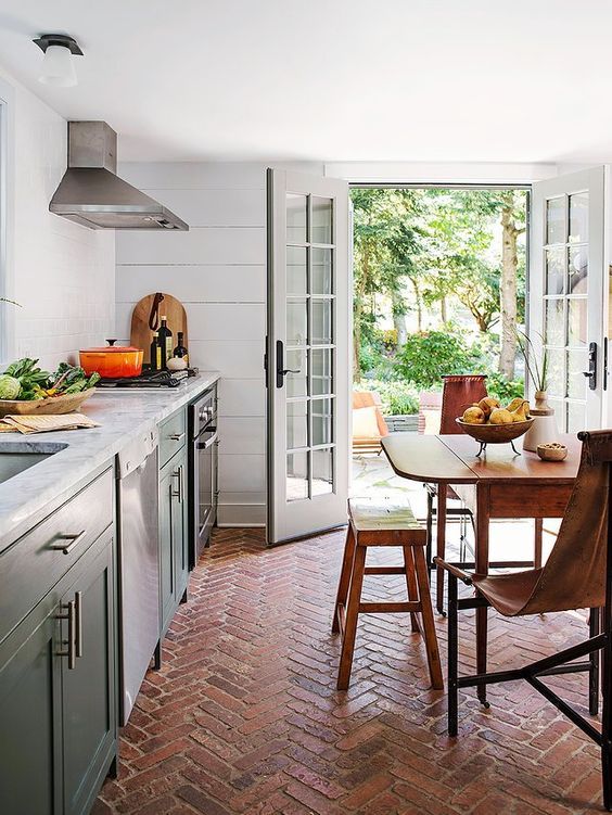 a farmhouse kitchen with green shaker cabinets with white stone countertops, a terracotta tile floor, a stained vintage table and chairs