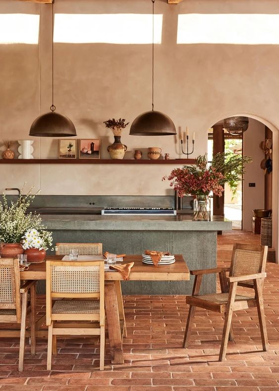 a farmhouse kitchen with tan plaster walls, a terracotta floor, a concrete kitchen island, a wooden dining set and metal pendant lamps