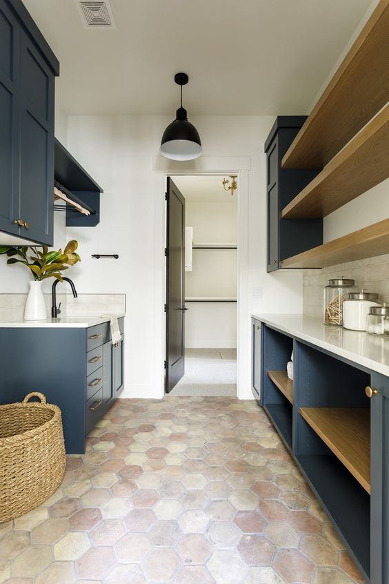 a farmhouse laundry with a terracotta tile floor, navy cabinets, white countertops, open shelves and a basket is very cozy