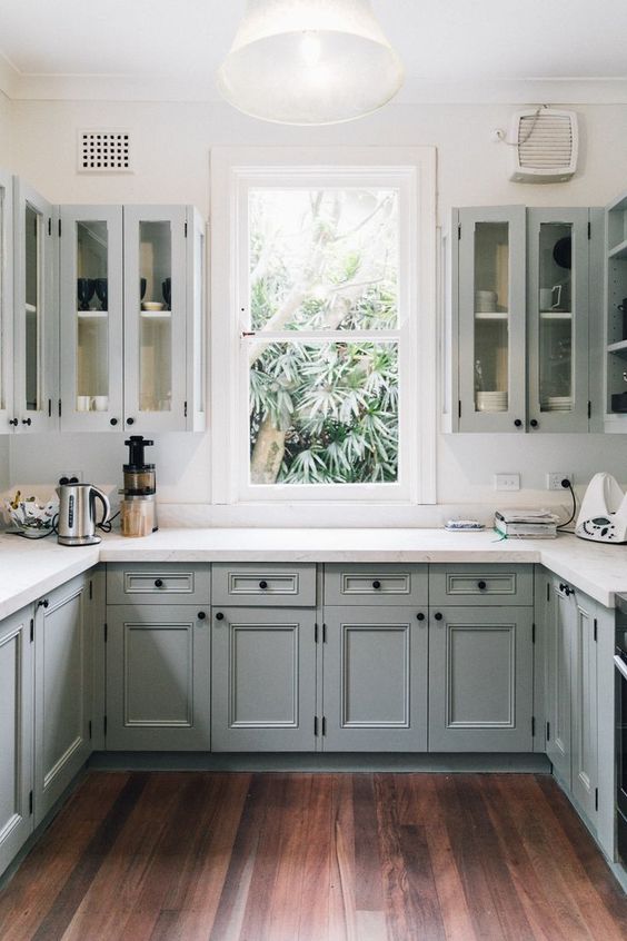 a light grey U-shaped kitchen with inlay and glass cabinets, white stone countertops and black knobs plus a window in the center