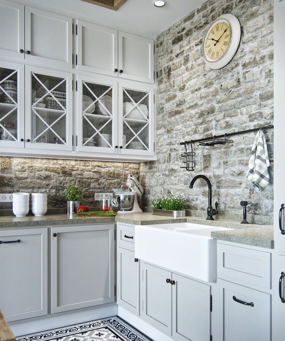 a light grey kitchen with inlay and glass front cabinets, a faux stone backsplash, grey granite countertops and black handles and fixtures