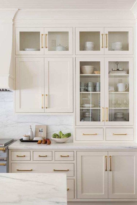 خزائن مطبخ زجاجية A-lovely-creamy-kitchen-with-shaker-and-glass-cabinets-white-marble-countertops-and-a-backsplash-and-gold-handles