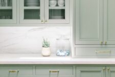 a lovely mint kitchen with shaker and glass front cabinets, a white marble backsplash and countertops and gold handles