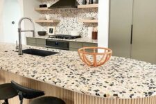 a lovely olive green kitchen with cheerful terrazzo countertops and a backsplash plus black stools and a hood