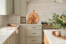 a lovely sage green kitchen with shaker cabinets, a white stone backsplash and countertops, a stained kitchen island and a chic pendant lamp