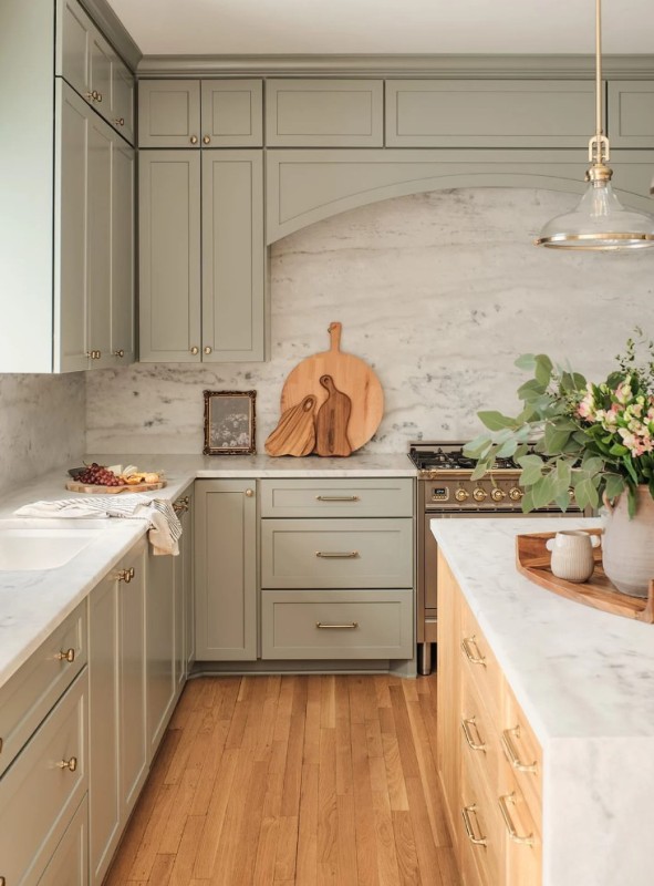 a lovely sage green kitchen with shaker cabinets, a white stone backsplash and countertops, a stained kitchen island and a chic pendant lamp