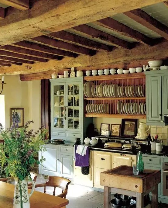 a lovely vintage kitchen with blue cabinets, a vintage hearth, open shelves for plates, wooden beams and a wooden dining set