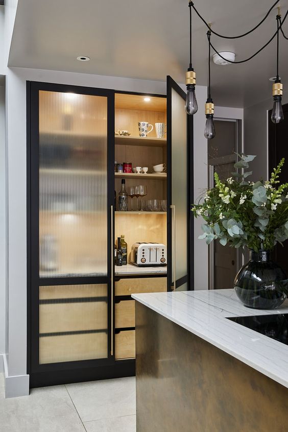 a mini built-in pantry with fluted glass black frame cabinets, black and gold pendant bulbs and a brass kitchen island