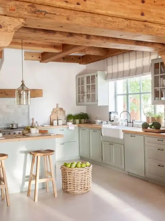 a modern cottage kitchen with wooden beams on the ceiling, light green shaker style cabinets, butcherblock countertops, a basket for storage and a pendant lamp