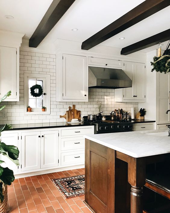 a modern farmhouse kitchen with a terracotta tile floor, a white subway tile backsplash, white shaker style cabinets, dark-stained wooden beams