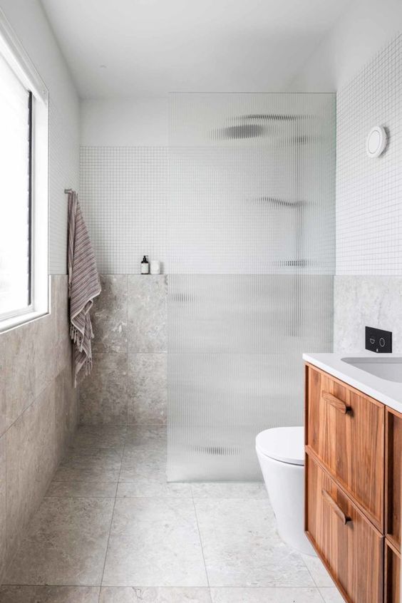 a modern laconic bathroom with neutral stone tiles, a shower space with a fluted glass space divider, a floating vanity and a window with much light