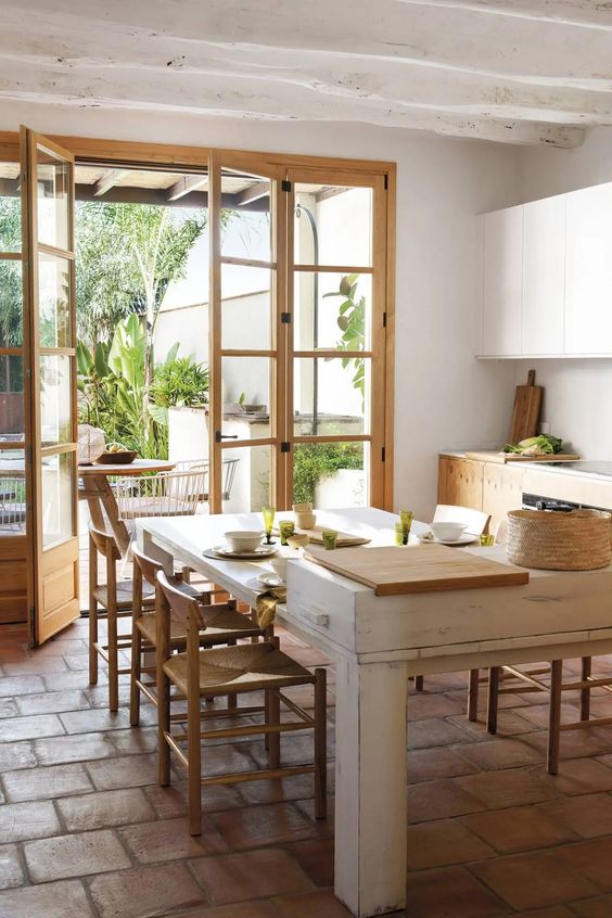 a modern rustic dining room with white walls and wooden beams, a terracotta tile floor, a whitewashed table and woven chairs