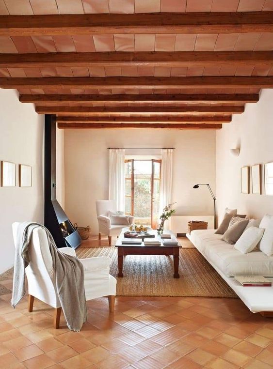 a modern rustic living room with a stained ceiling and wooden beams, a terracotta tile floor, white seating furniture, a black hearth
