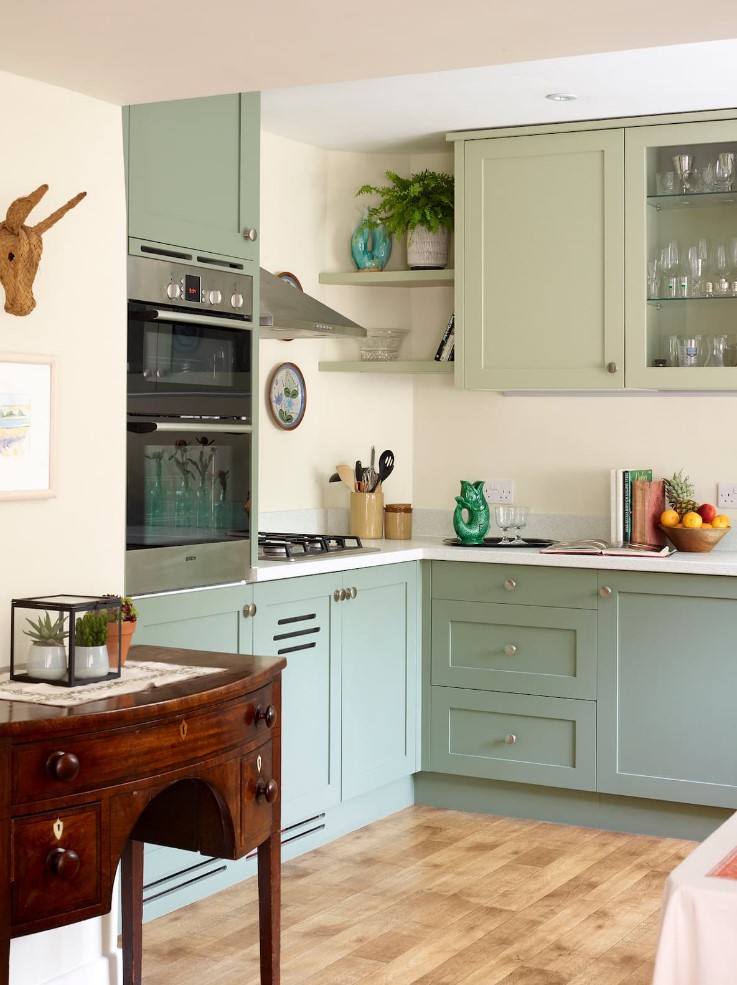 a modern sage green kitchen with shaker cabinets and glass front ones, a white backsplash and countertops plus a vintage sideboard