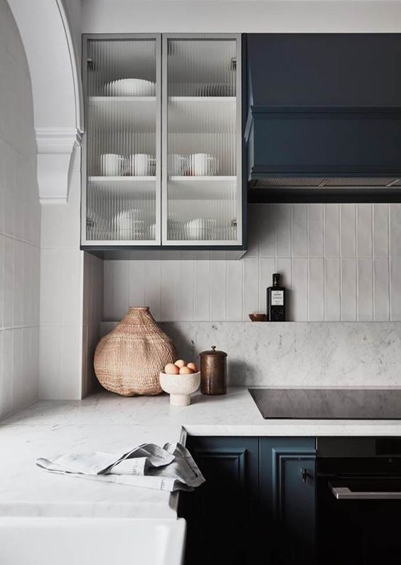 a navy kitchen with ornated and fluted glass cabinets, a white skinny tile backsplash and white stone countertops is a chic space