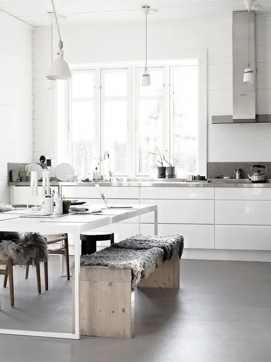 a neutral Nordic kitchen with sleke white cabinets, metal countertops, a white table and wooden benches plus pendant lamps