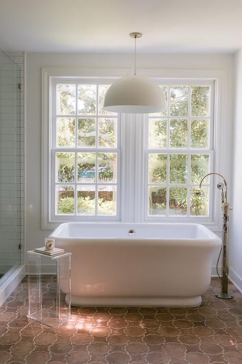 a neutral bathroom with a refined vintage feel, with a large window and a tub next to it, a terracotta tile floor and an acrylic stool