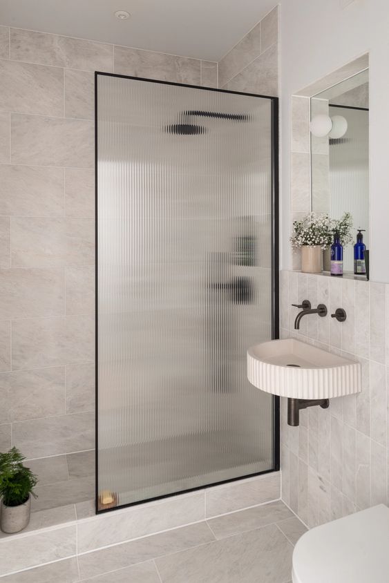 a neutral bathroom with stone tiles, a shower space with a fluted glass space divider, a wall-mounted sink and a mirror with a shelf