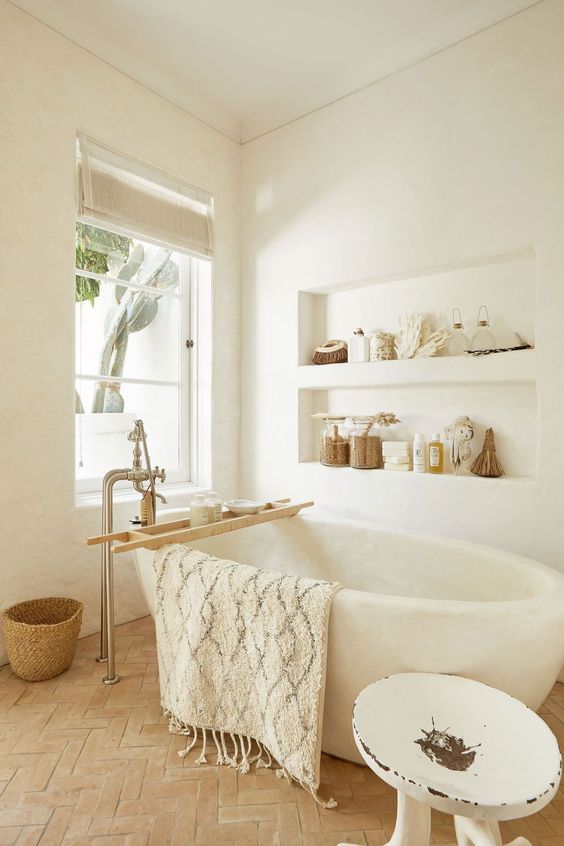 a neutral bathroom with white plaster walls with niches, a wabi-sabi tub, a terracotta floor, a white stool and baskets