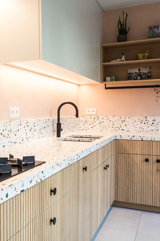a pastel kitchen with white and stained fluted cabinets, white terrazzo countertops and a backsplash, black fixtures