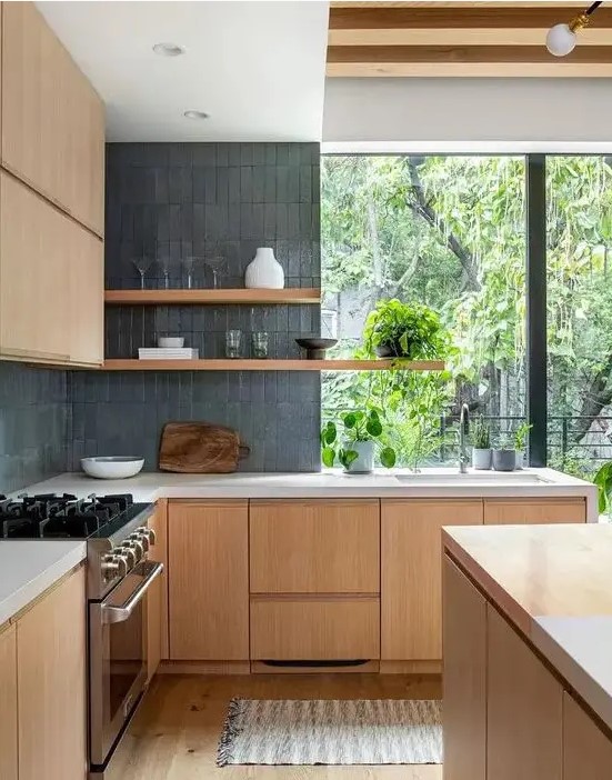 a pretty contemporary kitchen with sleek light-stained cabinetry, grey skinny tiles, open shelves and a large window with greenery views