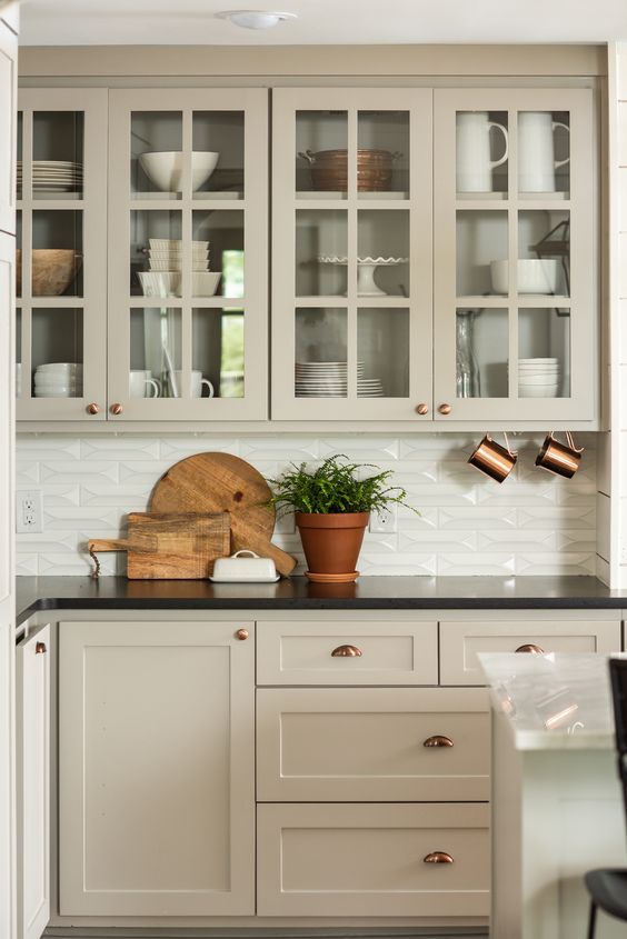 a refined greige kitchen with shaker and glass front cabinets, a white embossed tile backsplash and black countertops is a beautiful farmhouse solution