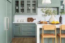 a sage green kitchen with shaker and glass front cabinets, white stone countertops and a backsplash, woven stools