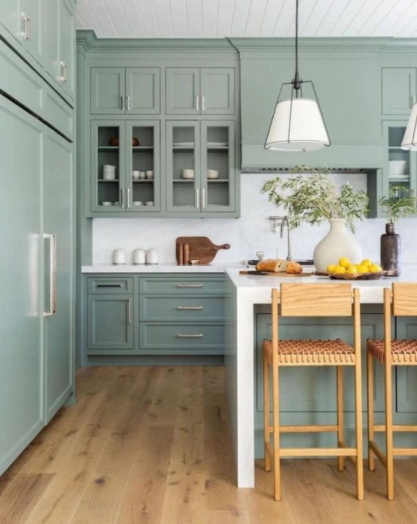 a sage green kitchen with shaker and glass front cabinets, white stone countertops and a backsplash, woven stools